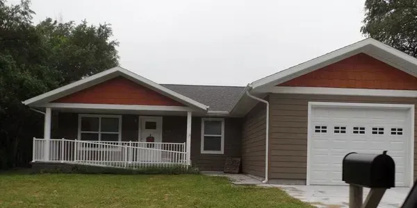 A brown house with a white porch and garage. It has a black mailbox, green grass, and overcast skies.