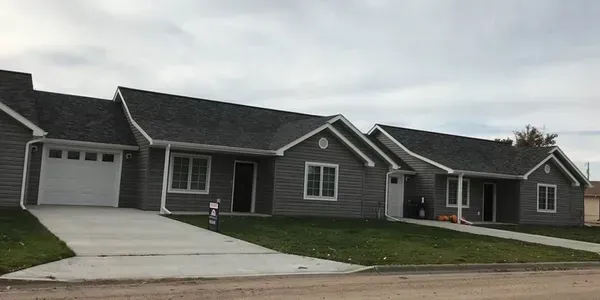 Gray houses with dark roofs under a cloudy sky. Driveways and green lawns in front.