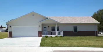 A beige house with a two-tone brick facade, white garage door, and a grassy yard under a blue sky.