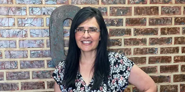 Woman with long dark hair, glasses, and floral top smiles in front of a brick wall and a large metal letter "Q".