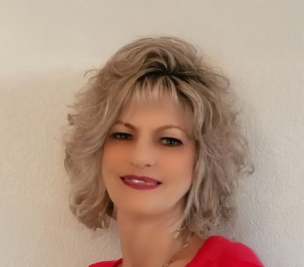 Woman with shoulder-length blonde curly hair smiles, wearing a red top. White wall background.