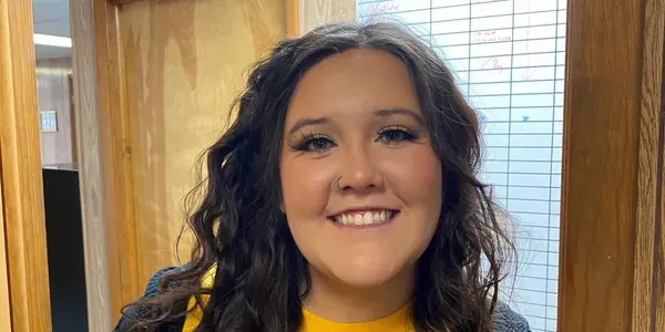 Woman with wavy dark hair smiles at the camera, wearing a yellow shirt and a grey sweater; inside a building.
