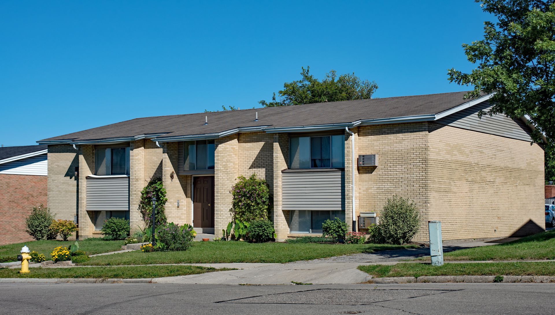 Two-story tan brick apartment building with dark roof, windows with blinds, and small front yard under a blue sky.
