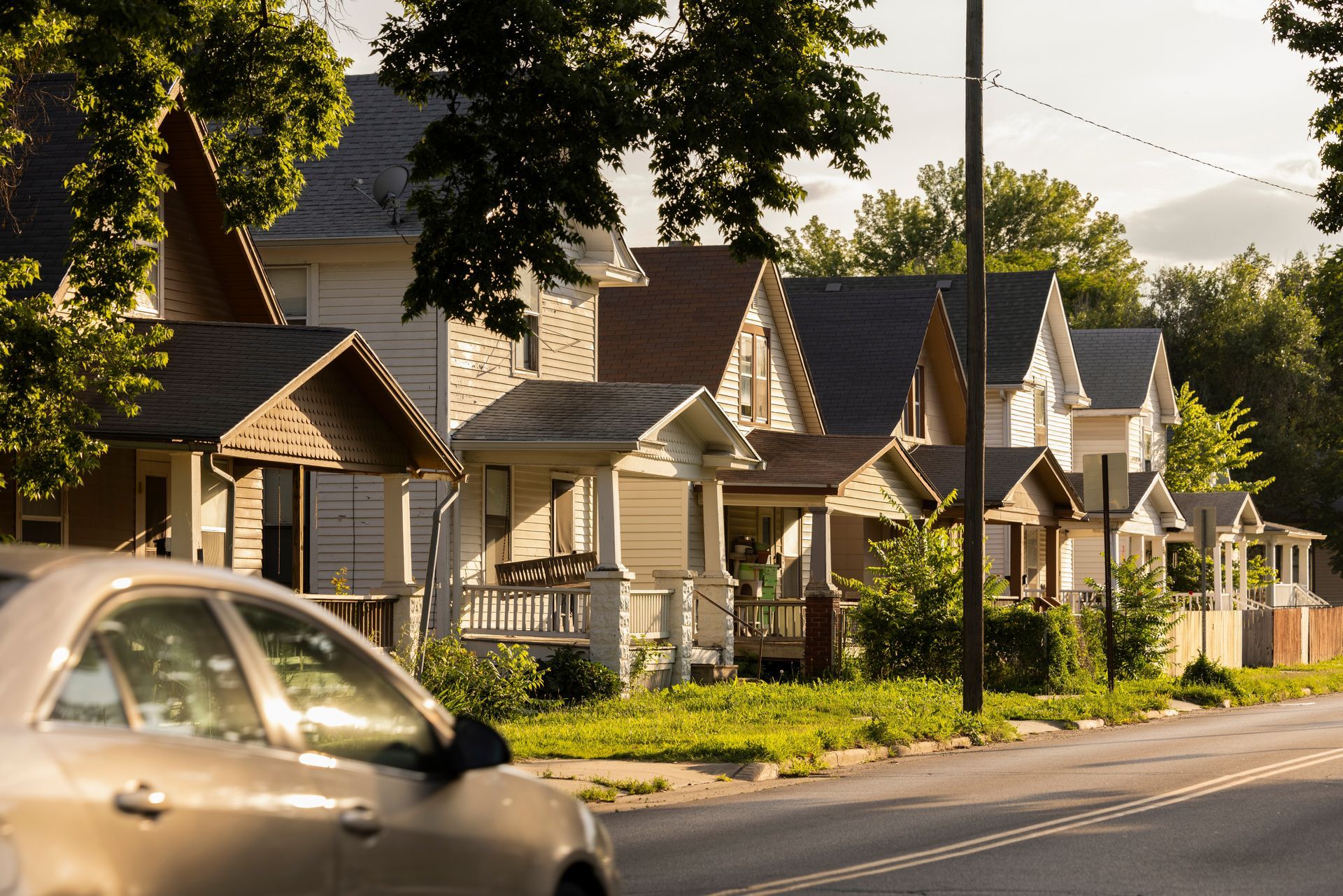 Row of houses with porches and trees along a sunny street; a car passes in the foreground.