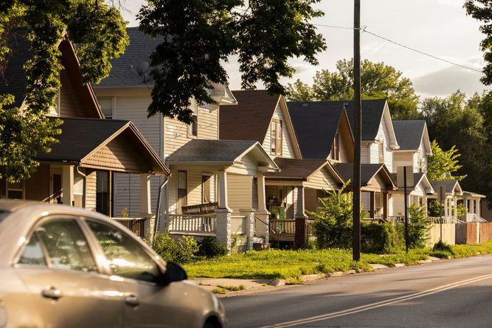 Row of houses with porches and trees along a sunny street; a car passes in the foreground.