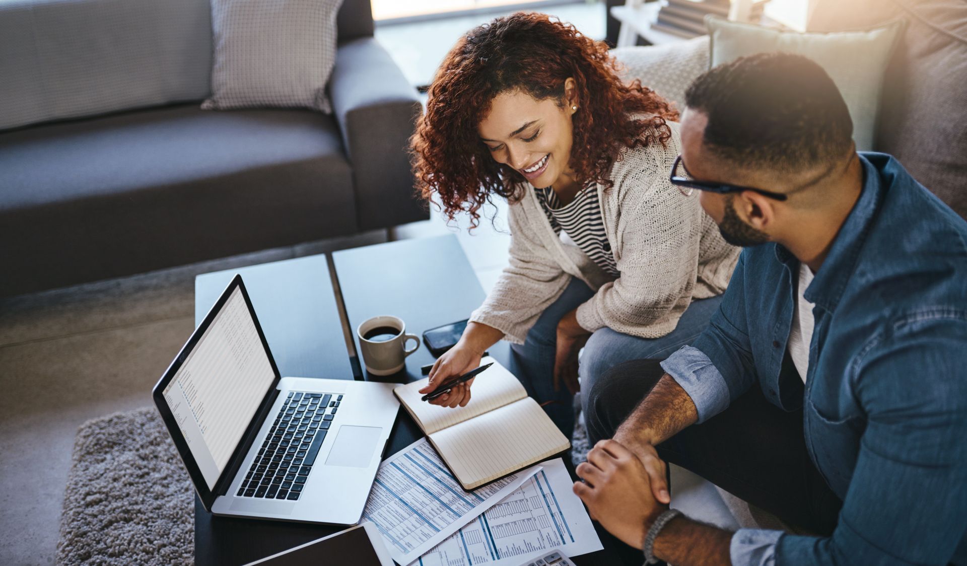 Couple reviews finances together at a coffee table with laptop, notebook, and papers.