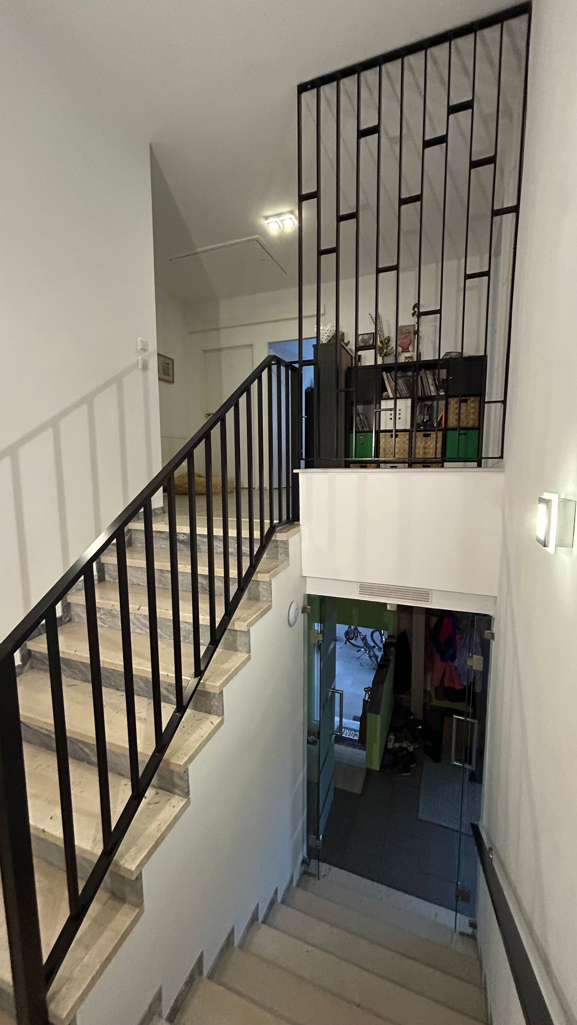 Treppenhaus mit weißen Wänden, schwarzem Geländer und dekorativer Metallwand. Ein Eingang im Untergeschoss ist sichtbar.