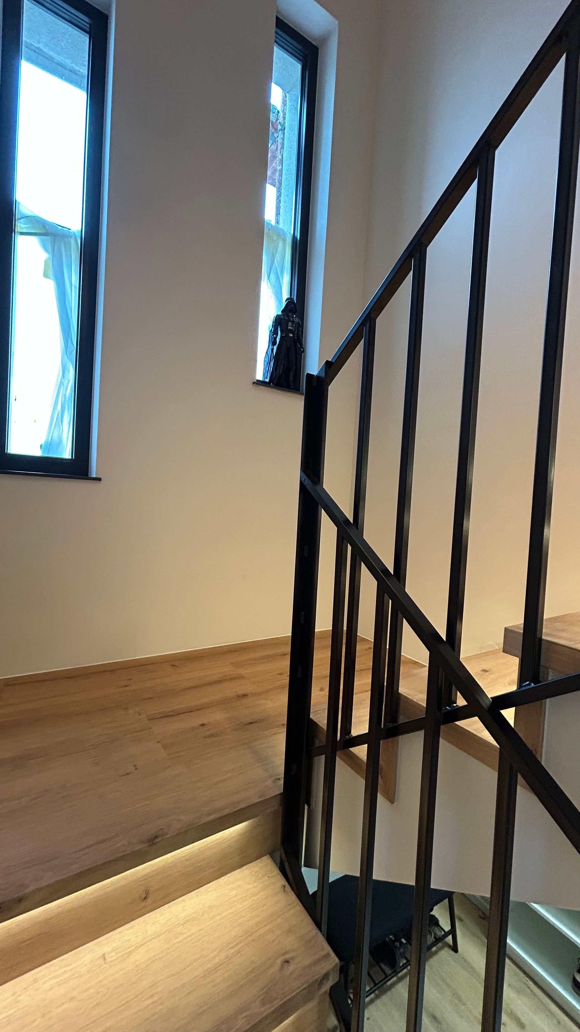 Holztreppe mit schwarzem Metallgeländer, in der Nähe von Fenstern, durch die Licht hereinströmt.