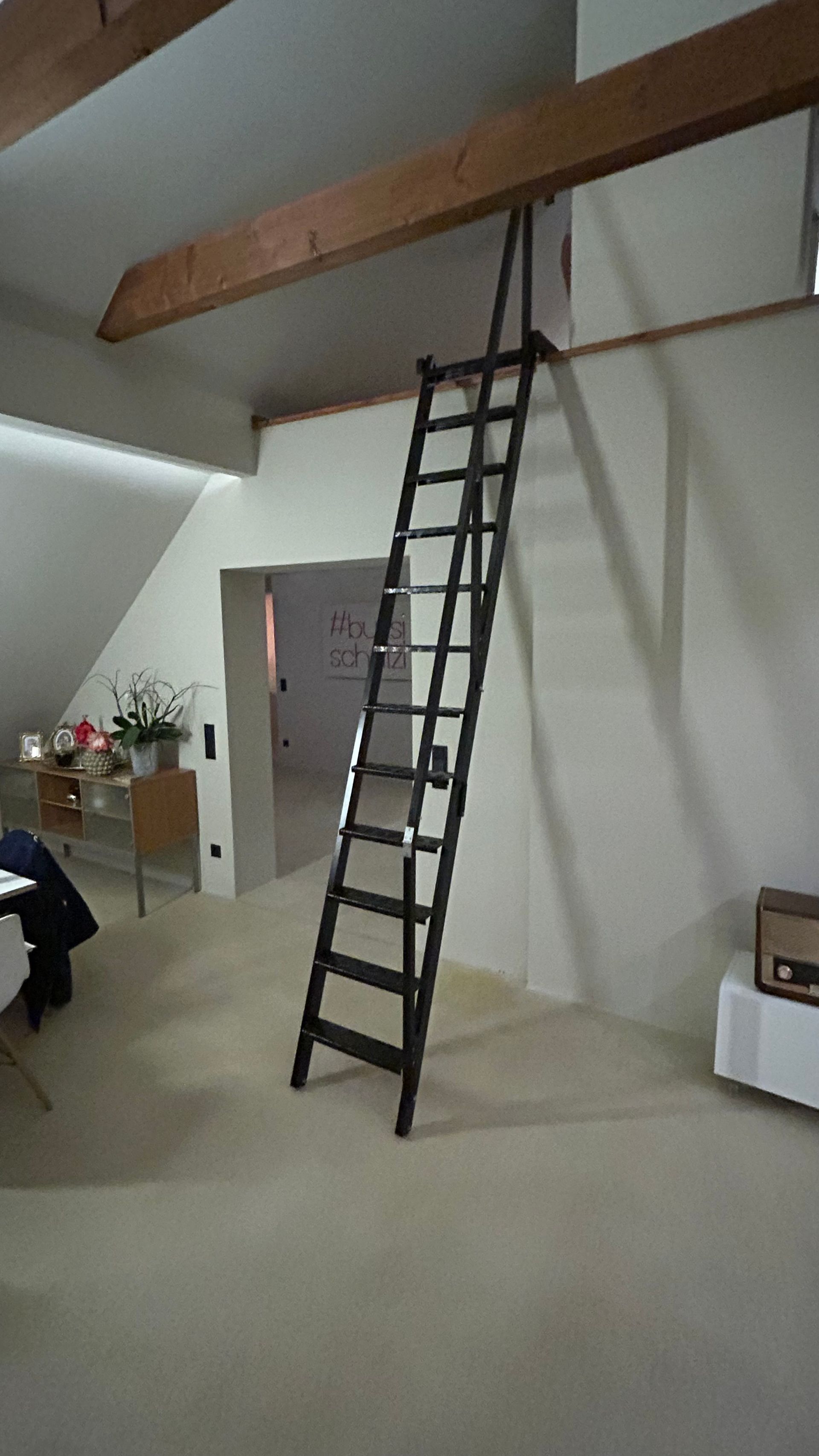 Eine schwarze Holzleiter lehnt an einer Wand und ist mit einem Deckenbalken in einem Raum verbunden.