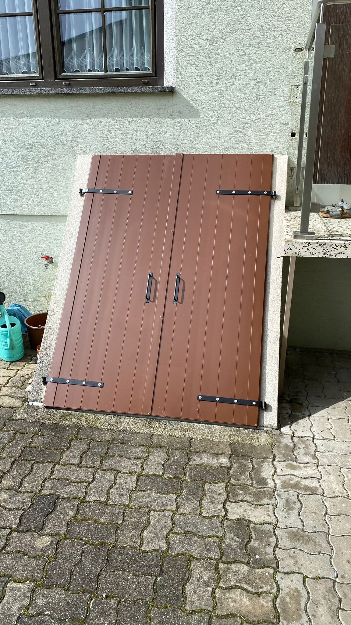 Auf einem gepflasterten Gehweg vor einem Haus steht ein braunes Tor mit Metall-Scharniere.