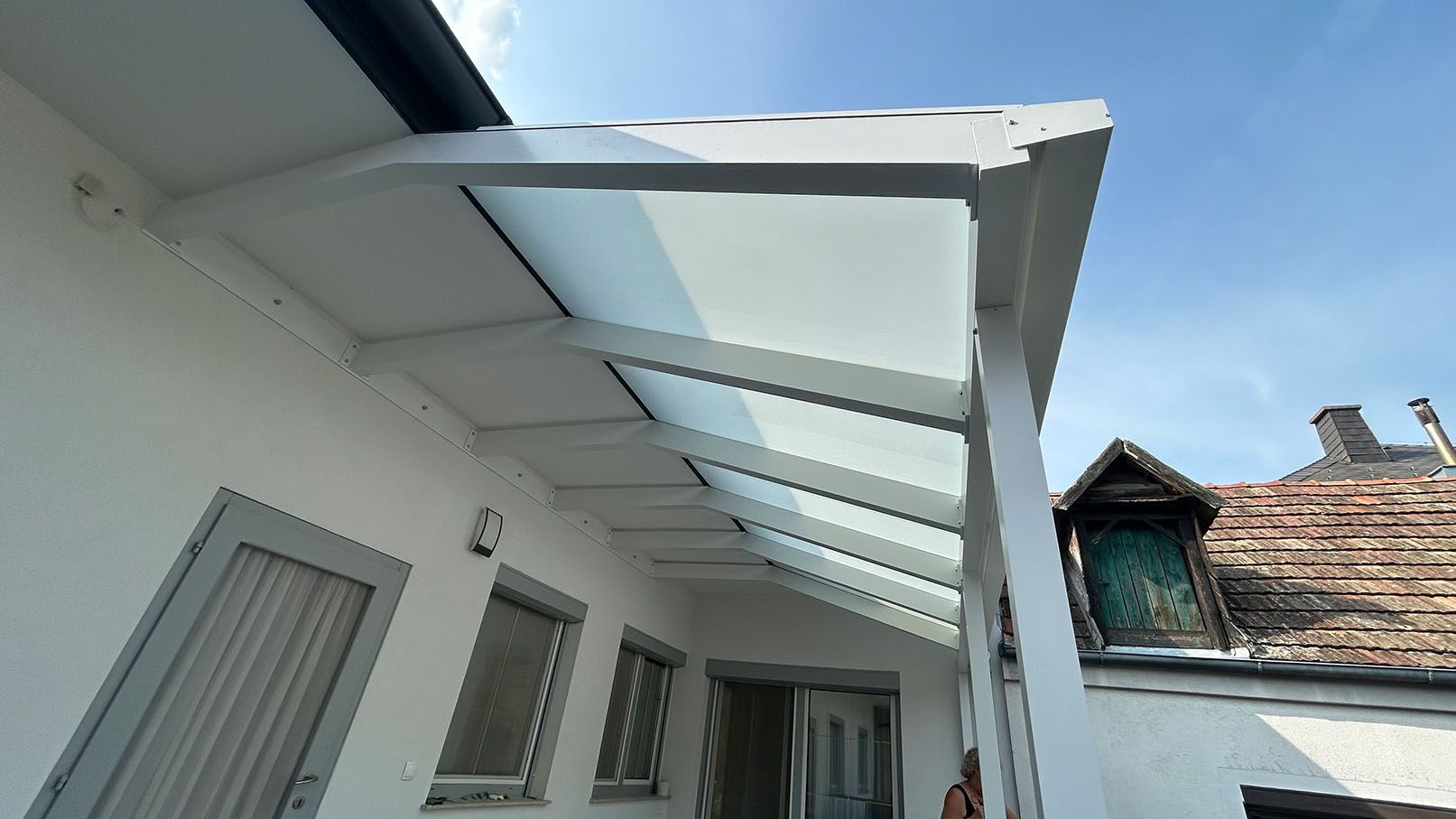 Weiße Markise aus Glas über einer Tür und Fenstern an der Seite eines weißen Gebäudes vor einem blauen Himmel.