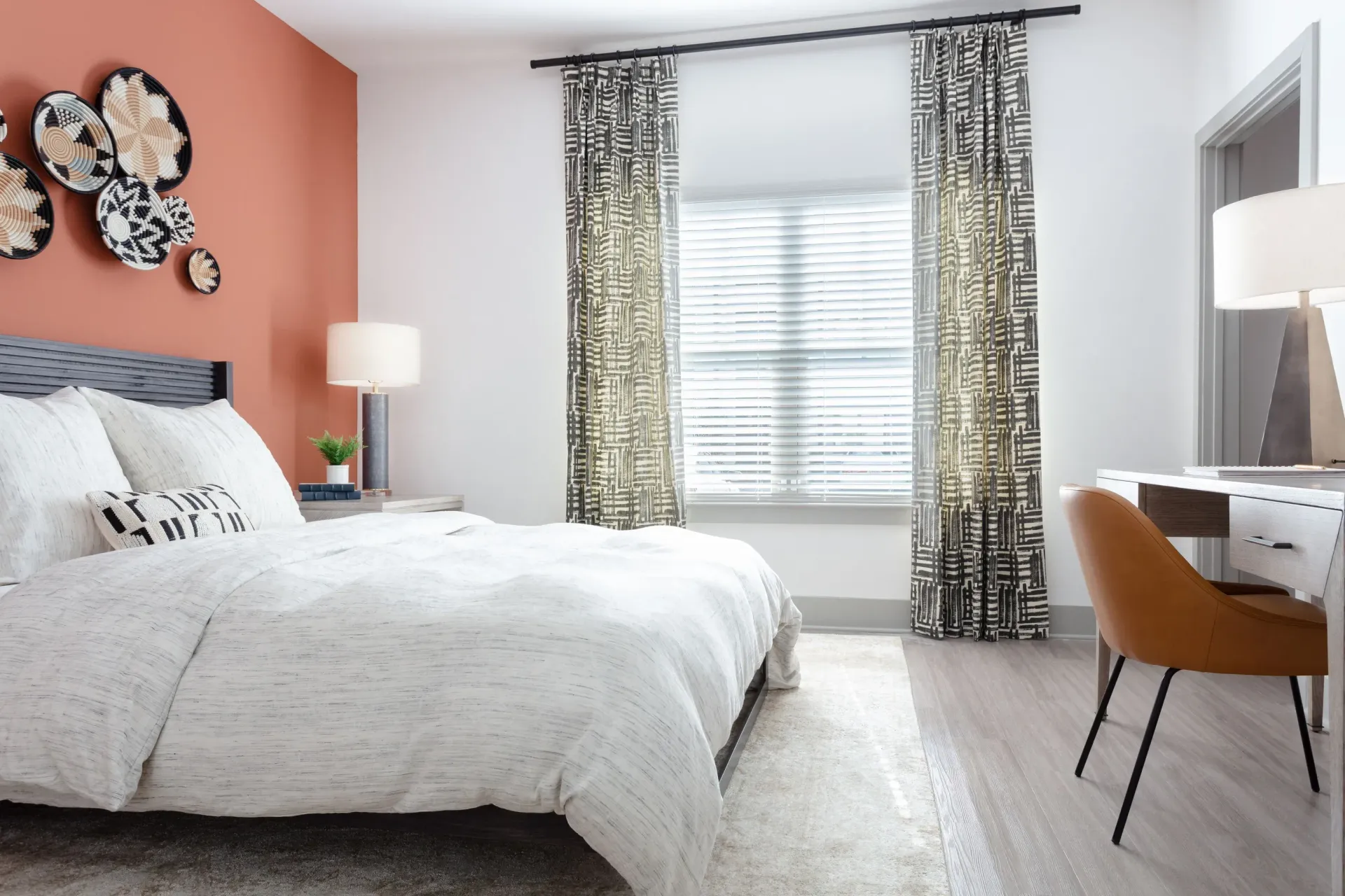Bedroom in a modern apartment with a white duvet, orange accent wall, nightstand lamp, desk, and patterned curtains.