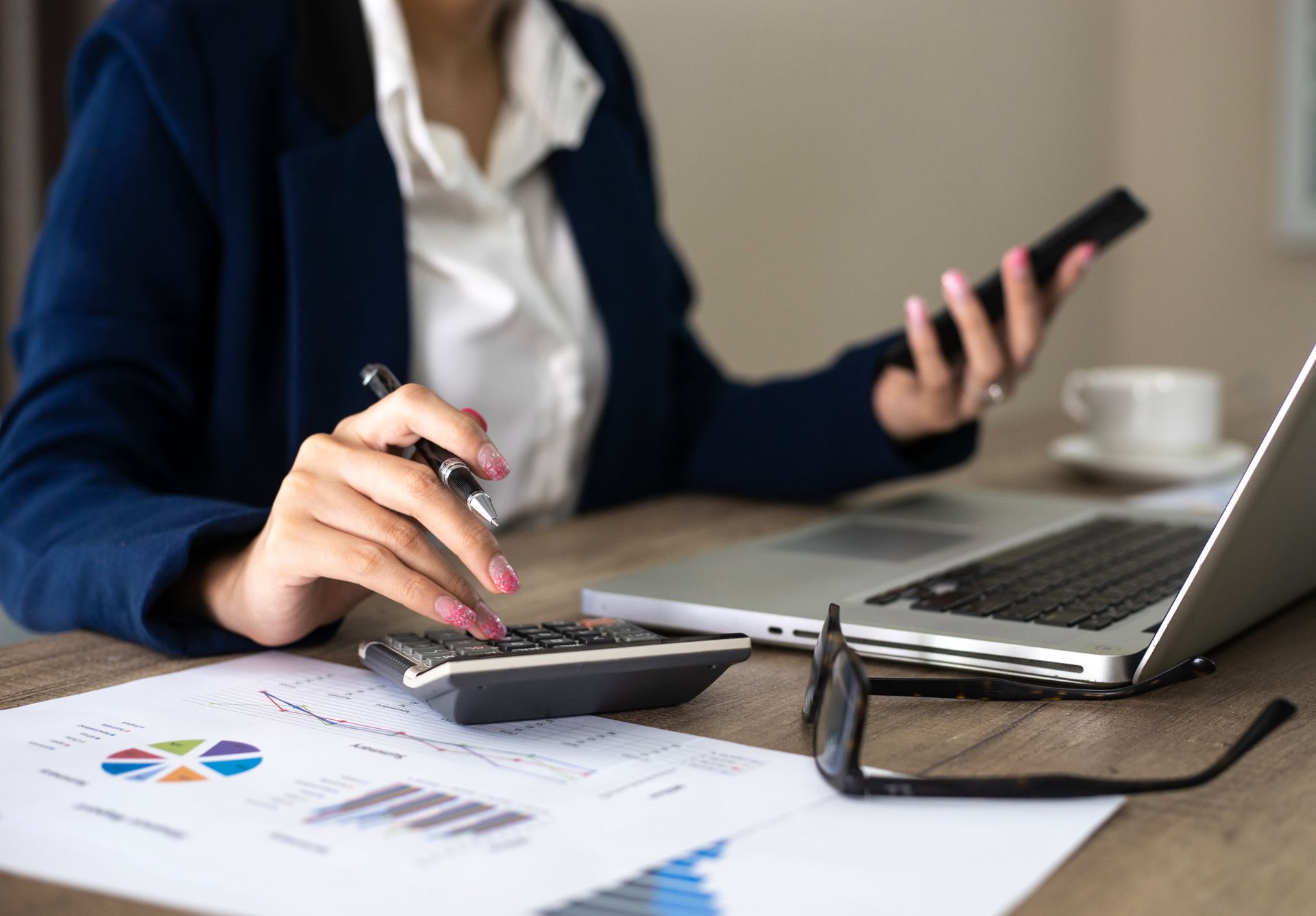 A woman is using a calculator and a laptop while holding a cell phone.