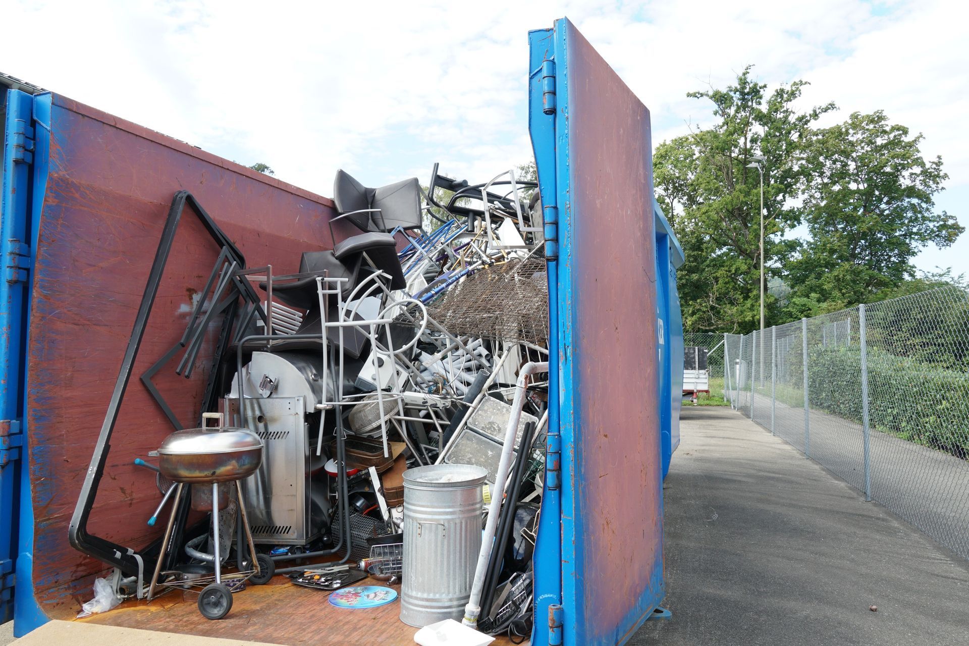 Blue and red dumpster overflowing with metal scrap, including chairs and a grill, outdoors.