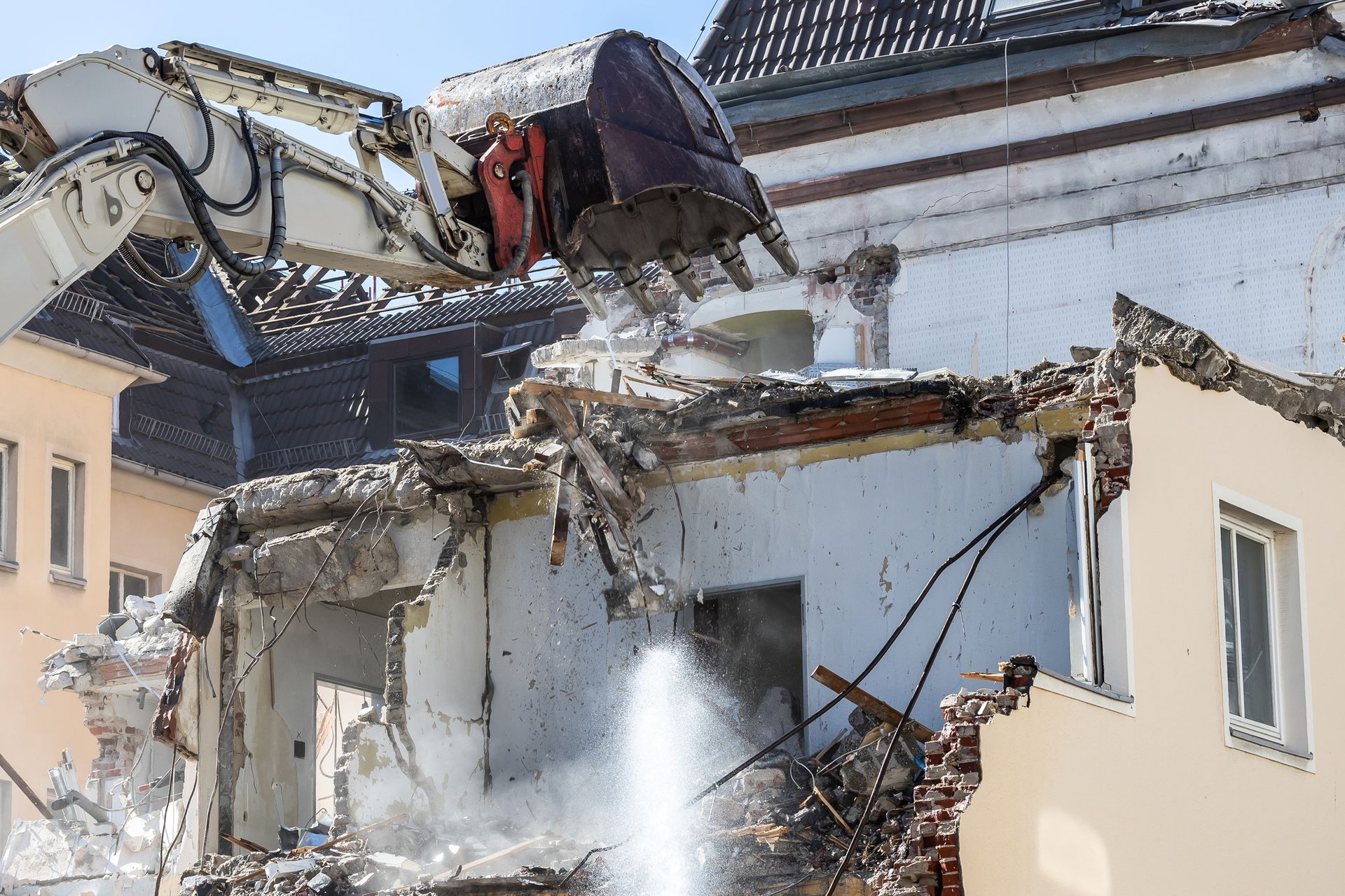 An excavator demolishing a building with falling debris and water spray to reduce dust.