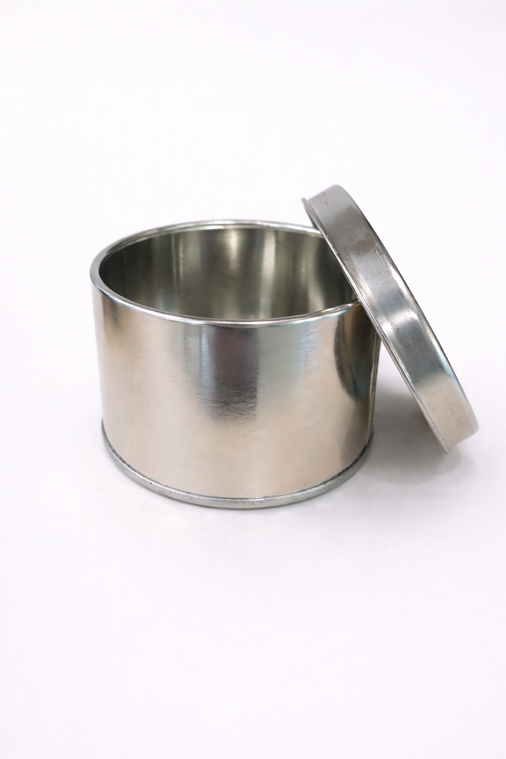 Pequeño recipiente de acero inoxidable con su tapa apoyada sobre un fondo blanco.