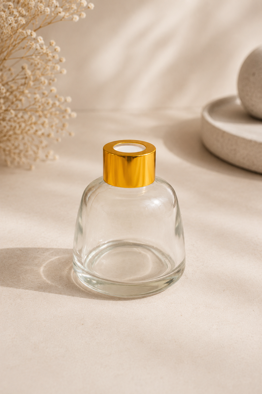Botella de vidrio transparente con tapón dorado sobre una superficie beige, con flores secas al fondo.