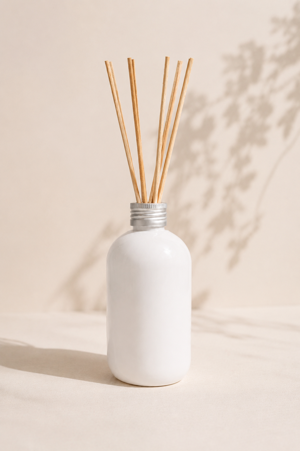Botella difusora de varillas blancas sobre una superficie beige iluminada por el sol con sombras en la pared.