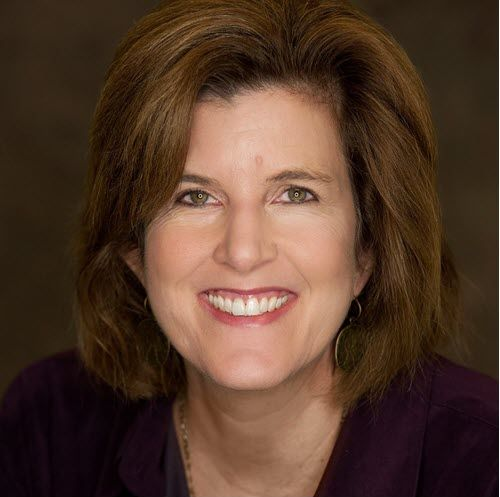 Woman with short brown hair smiles, wearing a purple top against a blurred background.
