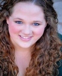 Woman with curly brown hair smiles; rosy cheeks, blue eyes, close-up shot.