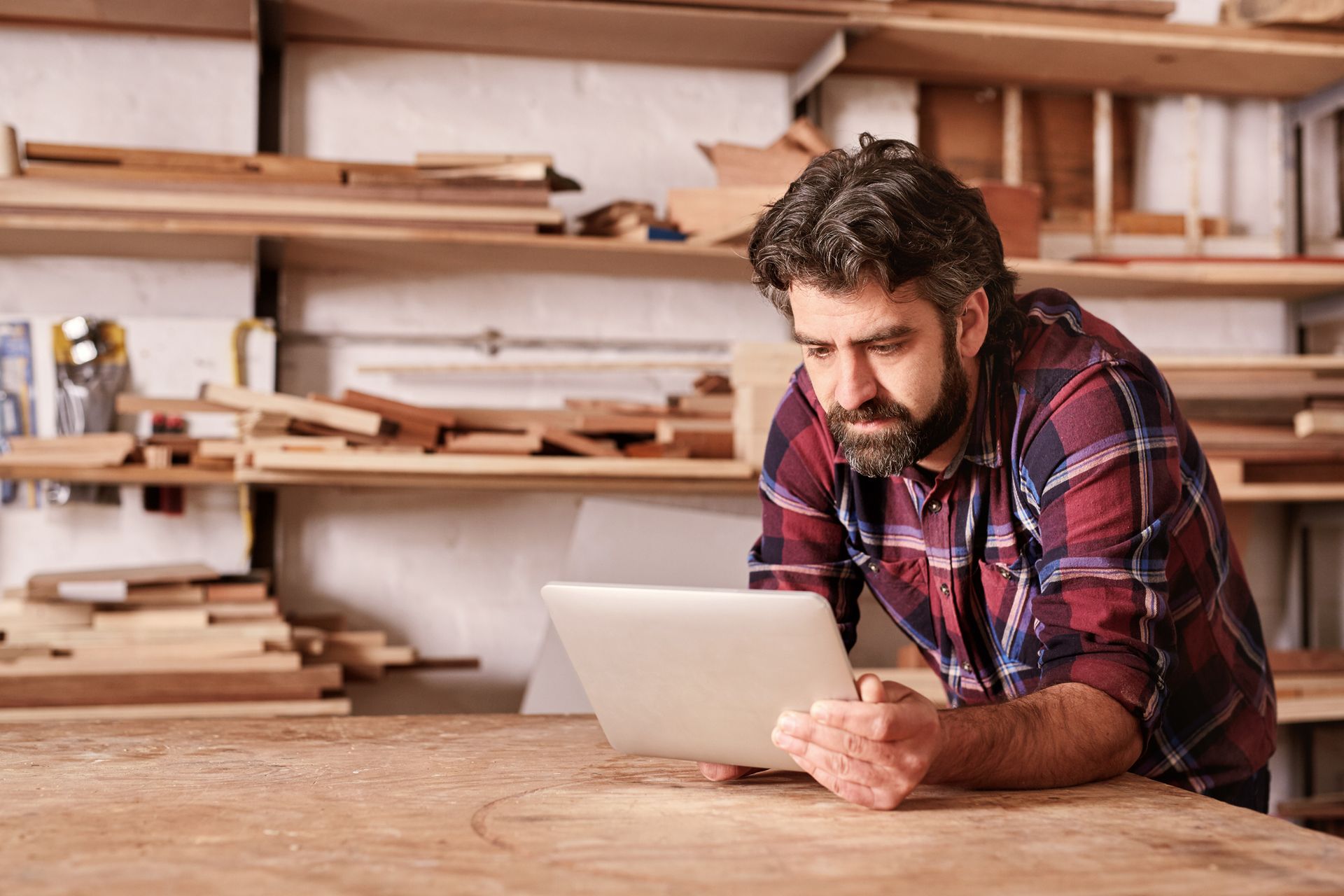 Woodworker in plaid shirt using a tablet at a workbench in a cluttered workshop.