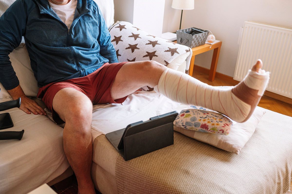 A person sits on a bed with their leg bandaged and elevated on a pillow, viewing a tablet.