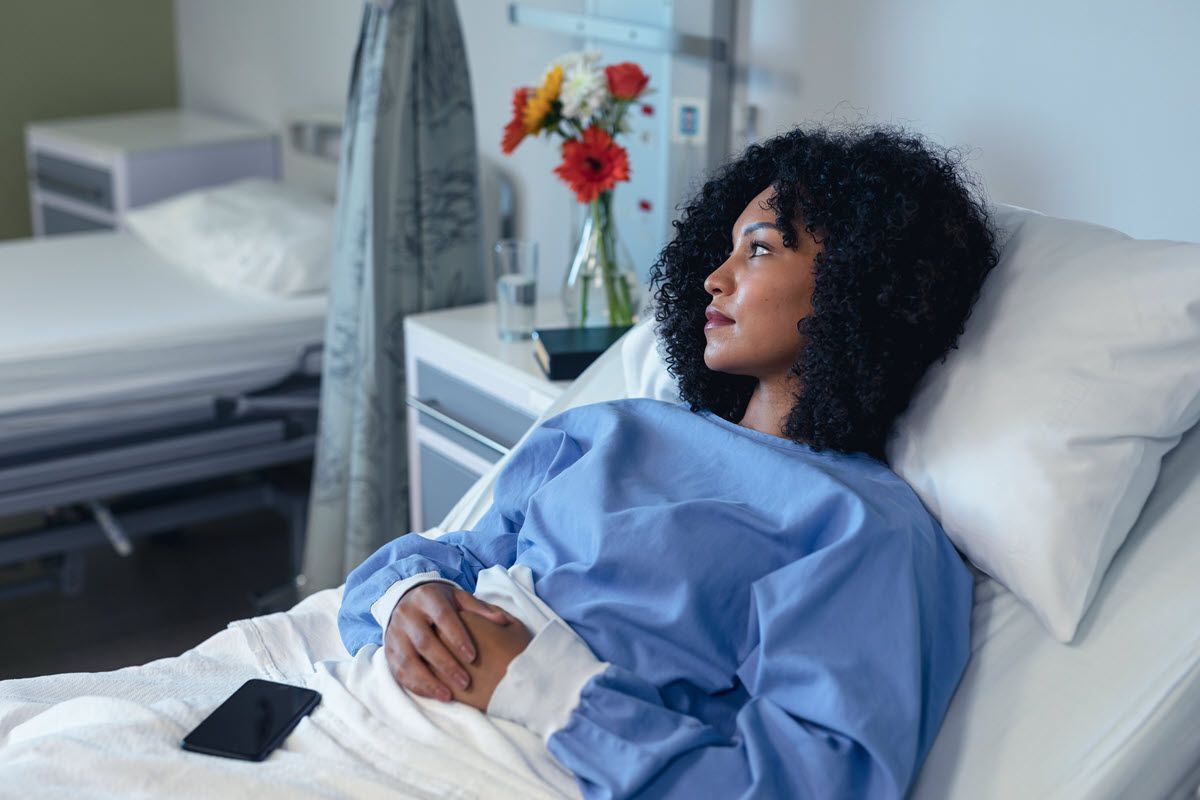 A person with curly dark hair wearing a blue hospital gown reclines in a hospital bed, looking away with a pensive gaze.