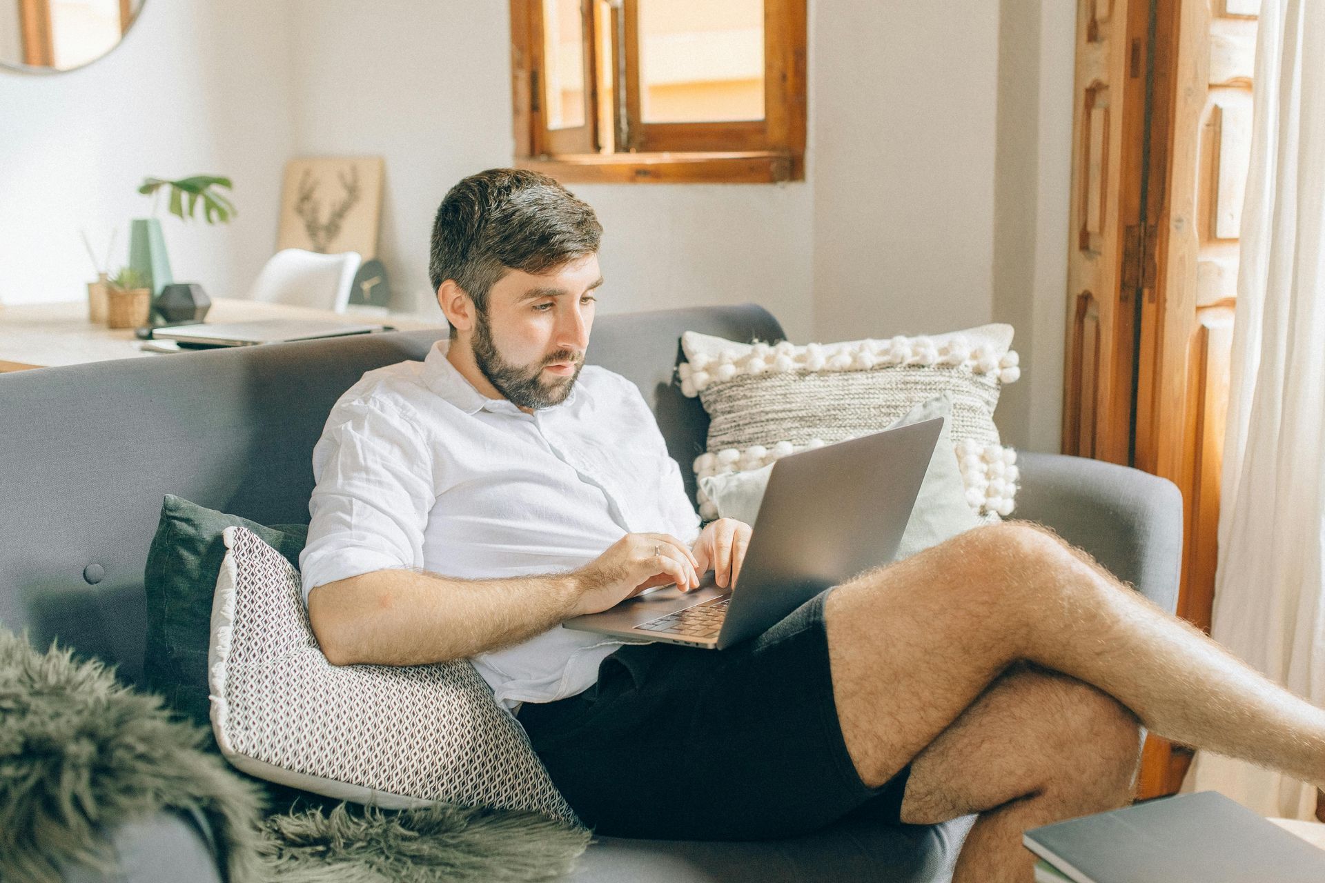 A person sitting on a sofa in a living room, wearing a white shirt and black shorts, working on a laptop.