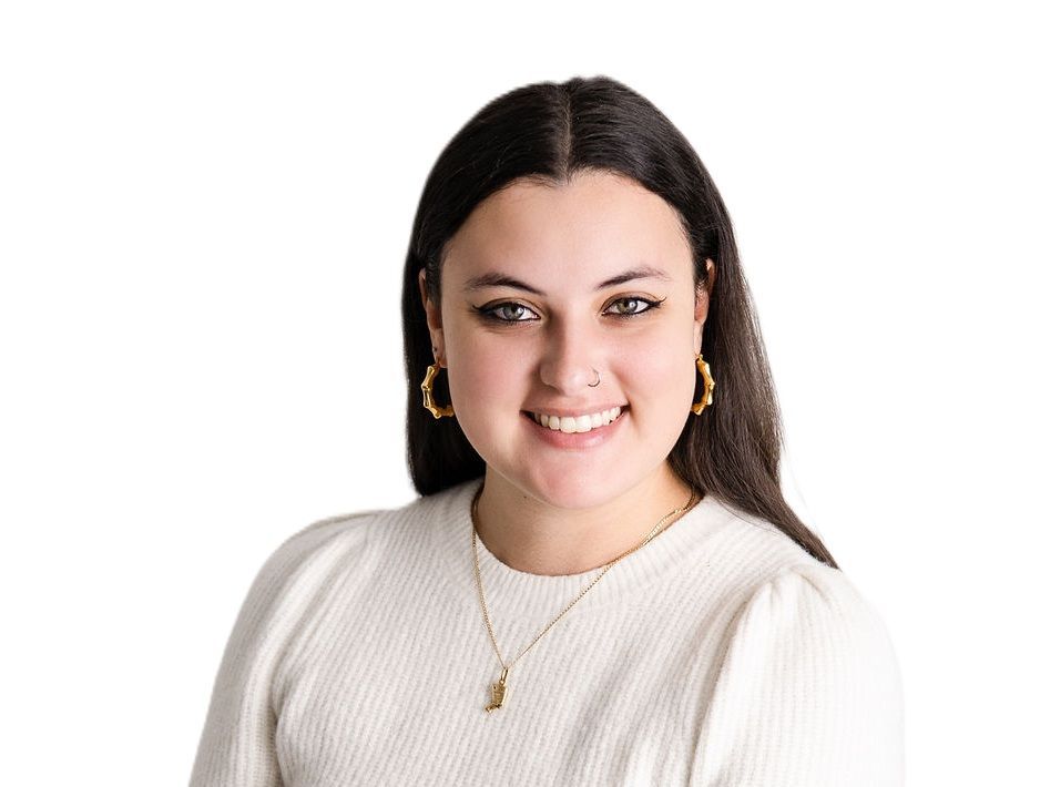 Tanna Beautement with dark hair wearing gold jewelry and a cream sweater against a white background.
