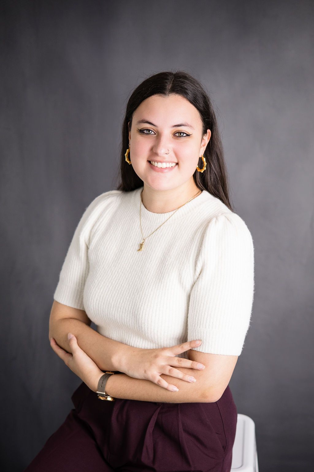 Tanna Beautement smiles, arms crossed, wearing a white sweater, gold jewelry, and maroon pants