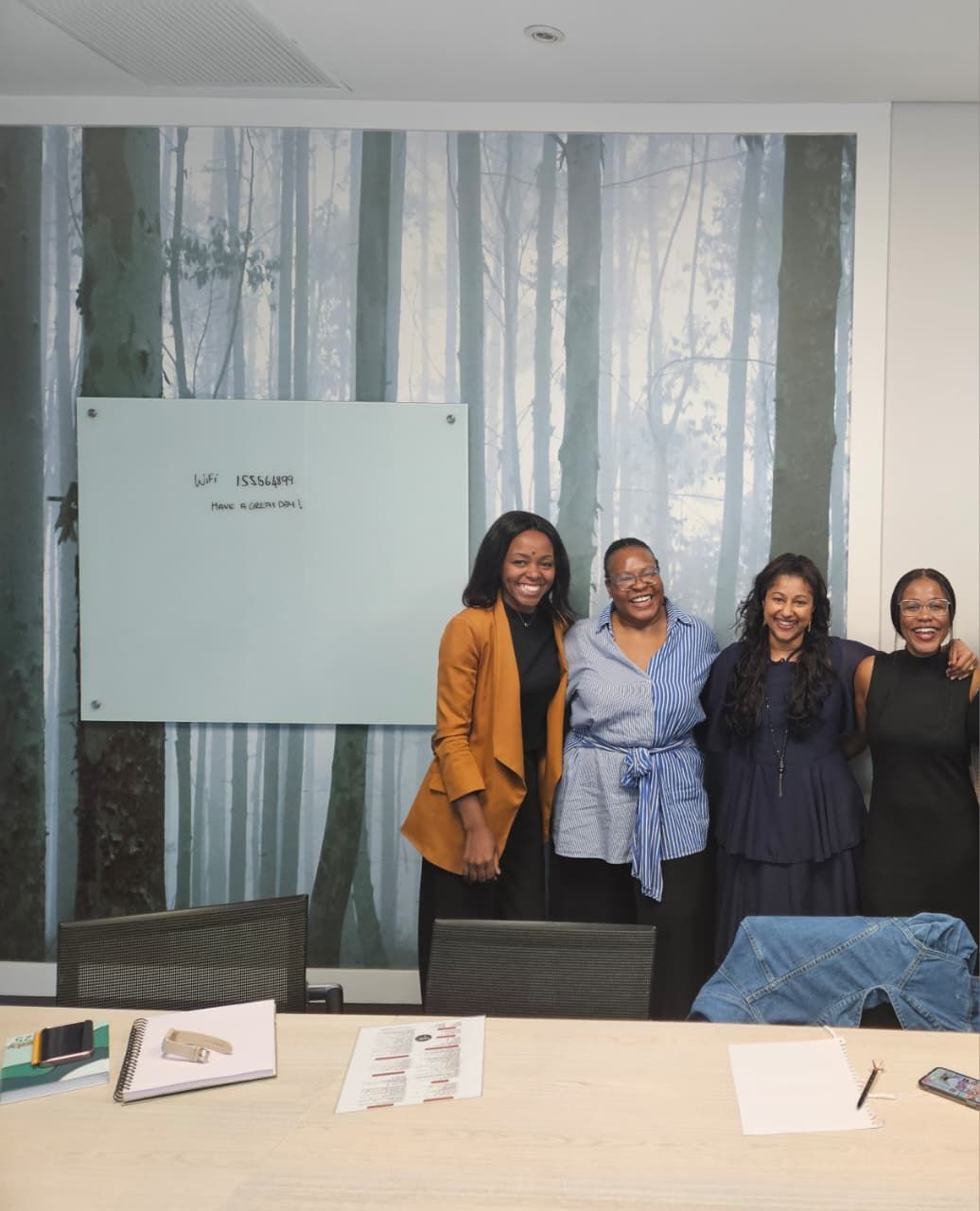 Sandy, Zinzisa, Phozisa and Tricia standing in a row and smiling in front of a whiteboard with text and a forest wall mural in an office.