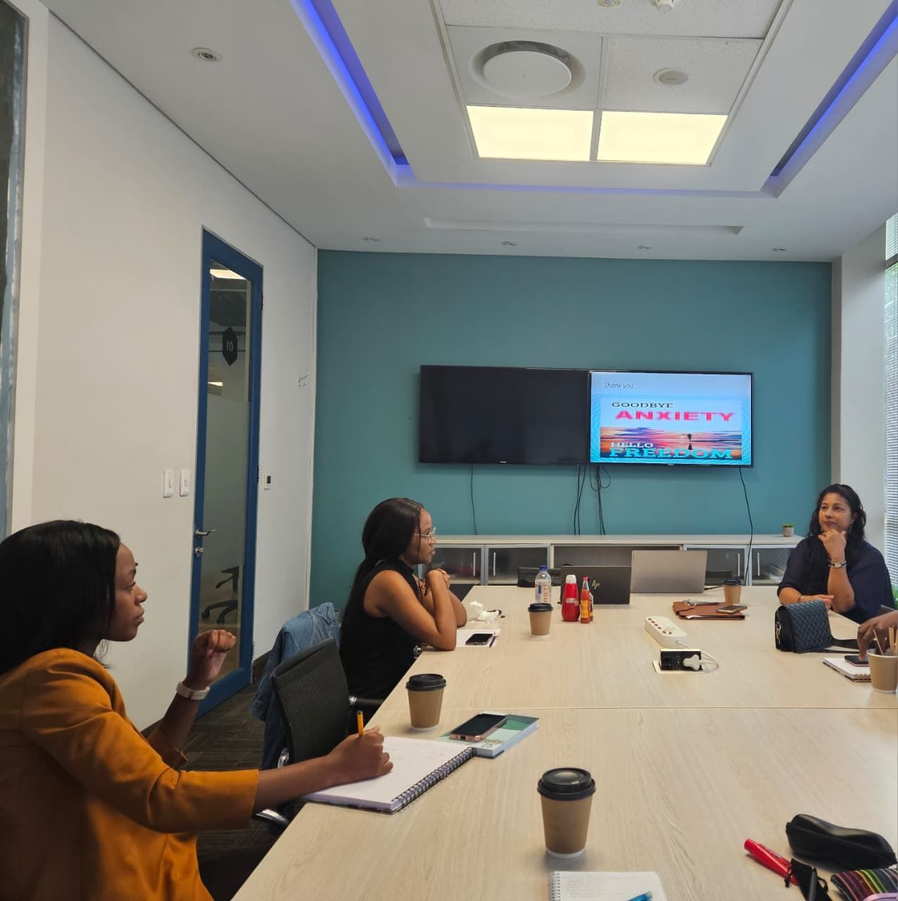 Zinzisa, Phozisa and Tricia sit at a conference table in a modern meeting room with a presentation on 