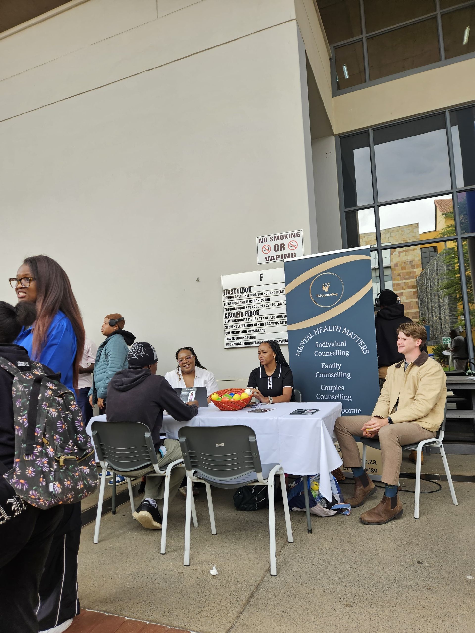 Students sit at a table outdoors under a tall TM Counselling promotional banner for a university event, with others standing nearby.