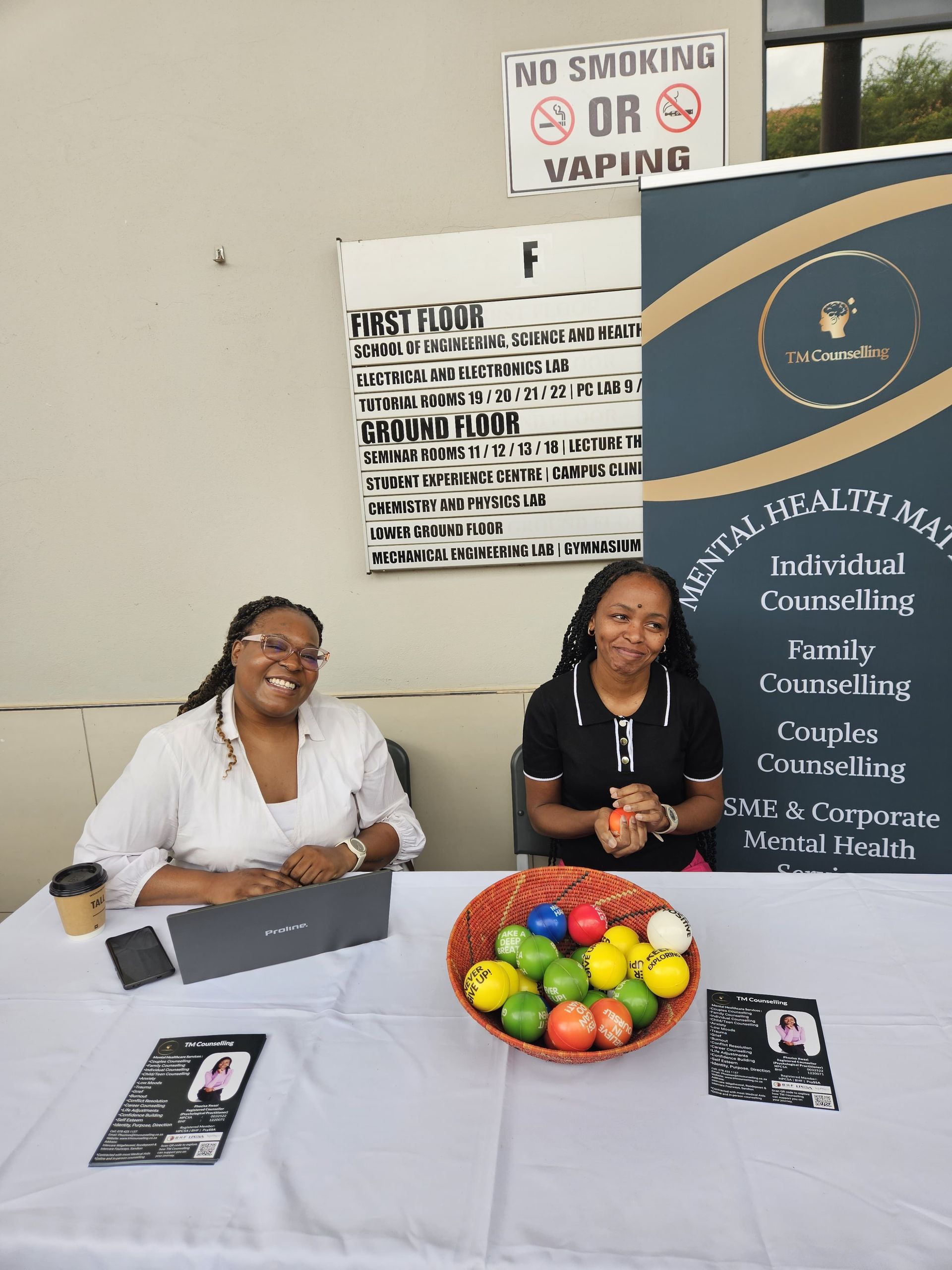 Sandy Kass and Phozisa Xwazi smile behind a table with a bowl of fruits, brochures, and a mental health service banner at an indoor event.