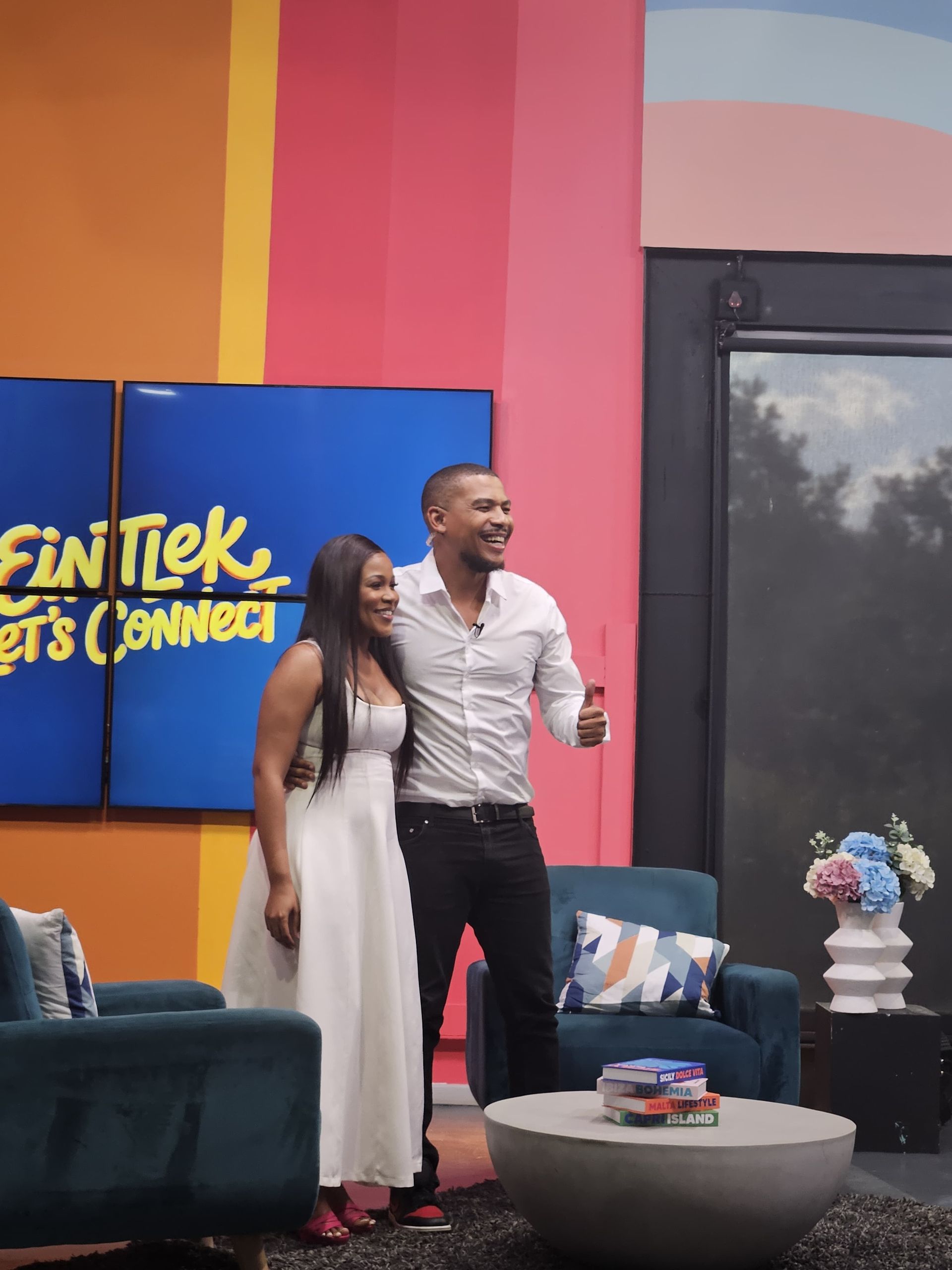 Zinzisa and Fran-Rico Lucas smiling  and stands together on a colorful television studio set with a blue 