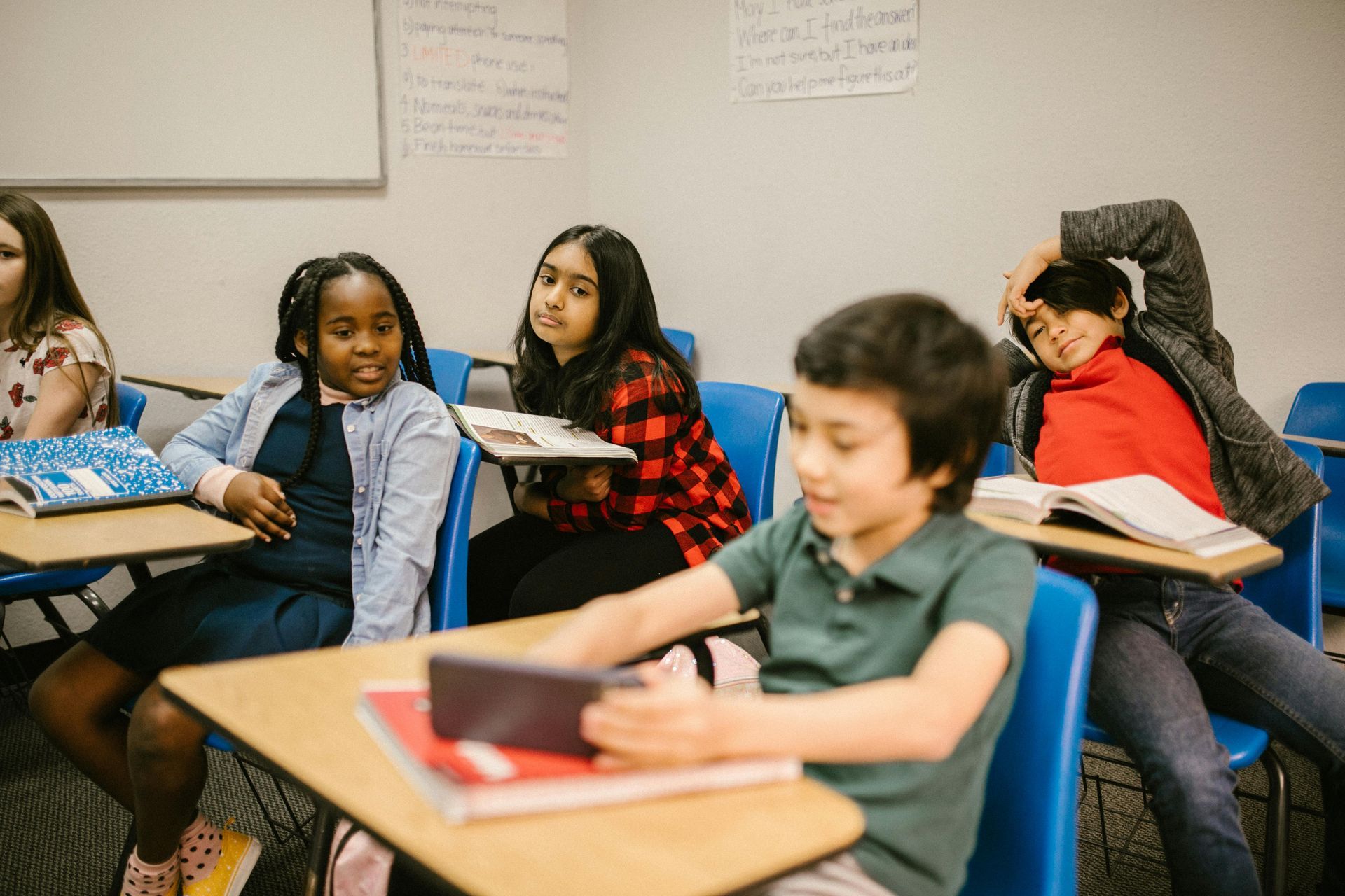 Children in a classroom seated at desks; some looking around, one using a tablet, and one with their hand on their head.