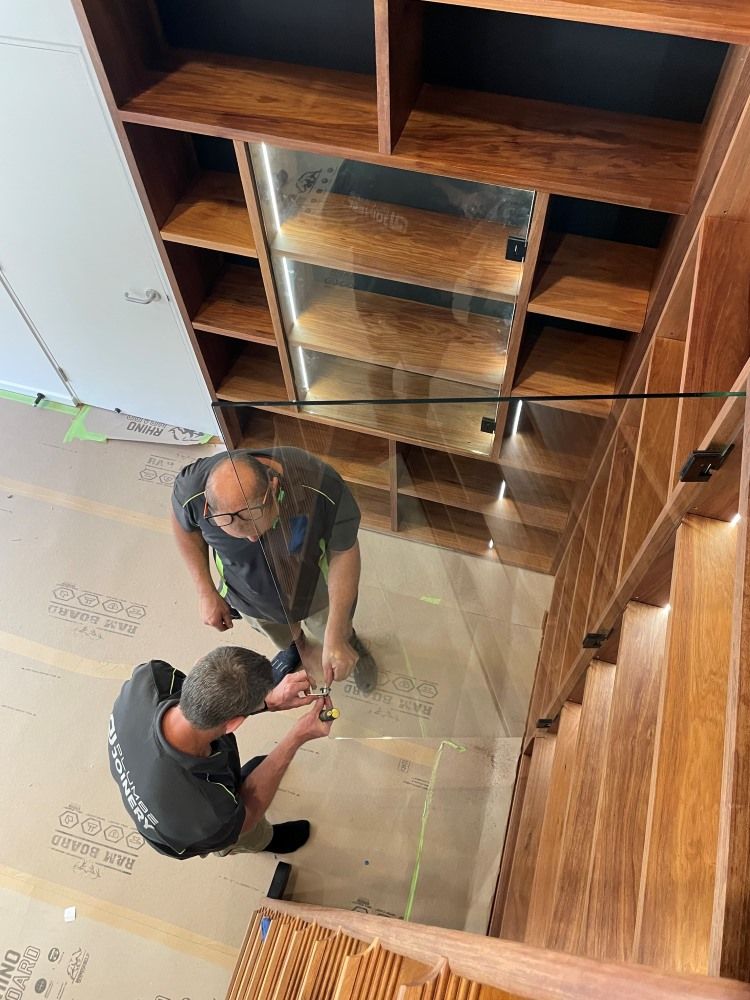 Two Workers Installing a Glass Panel Inside a Wooden Shelving Unit — Plumbe Joinery in Coffs Harbour, NSW