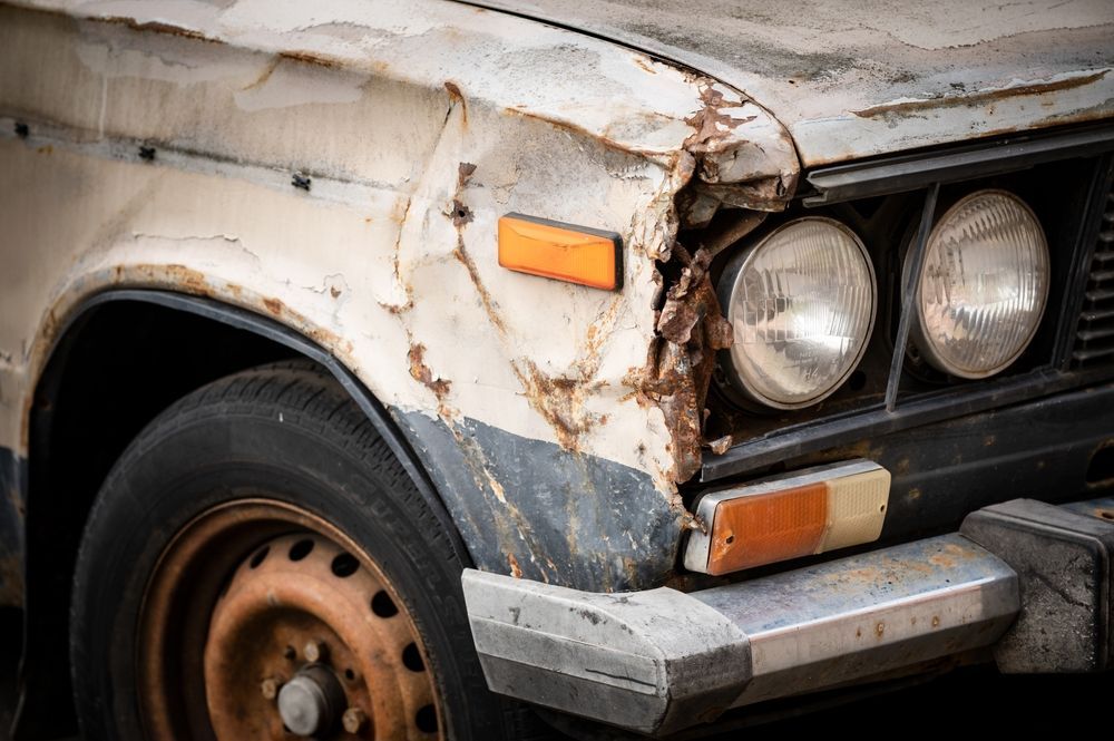 Damaged, weathered white car front; crushed fender, rusty wheel, cracked headlight.