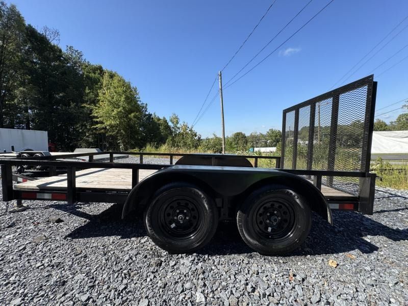 Black utility trailer with dual wheels, black fenders, and a mesh gate in a gravel lot on a sunny day.