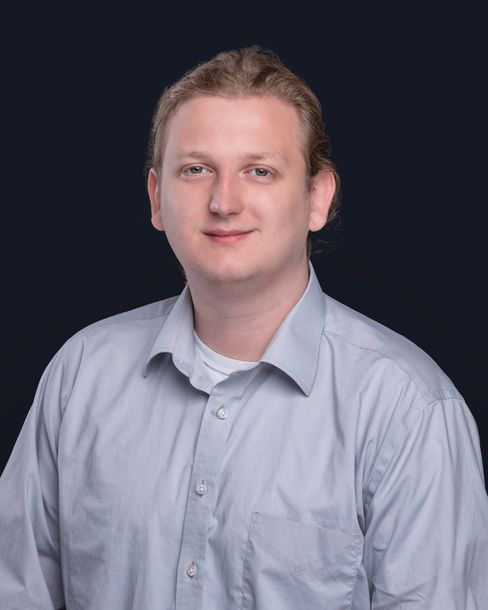 A man in a blue shirt is standing in front of a black background.