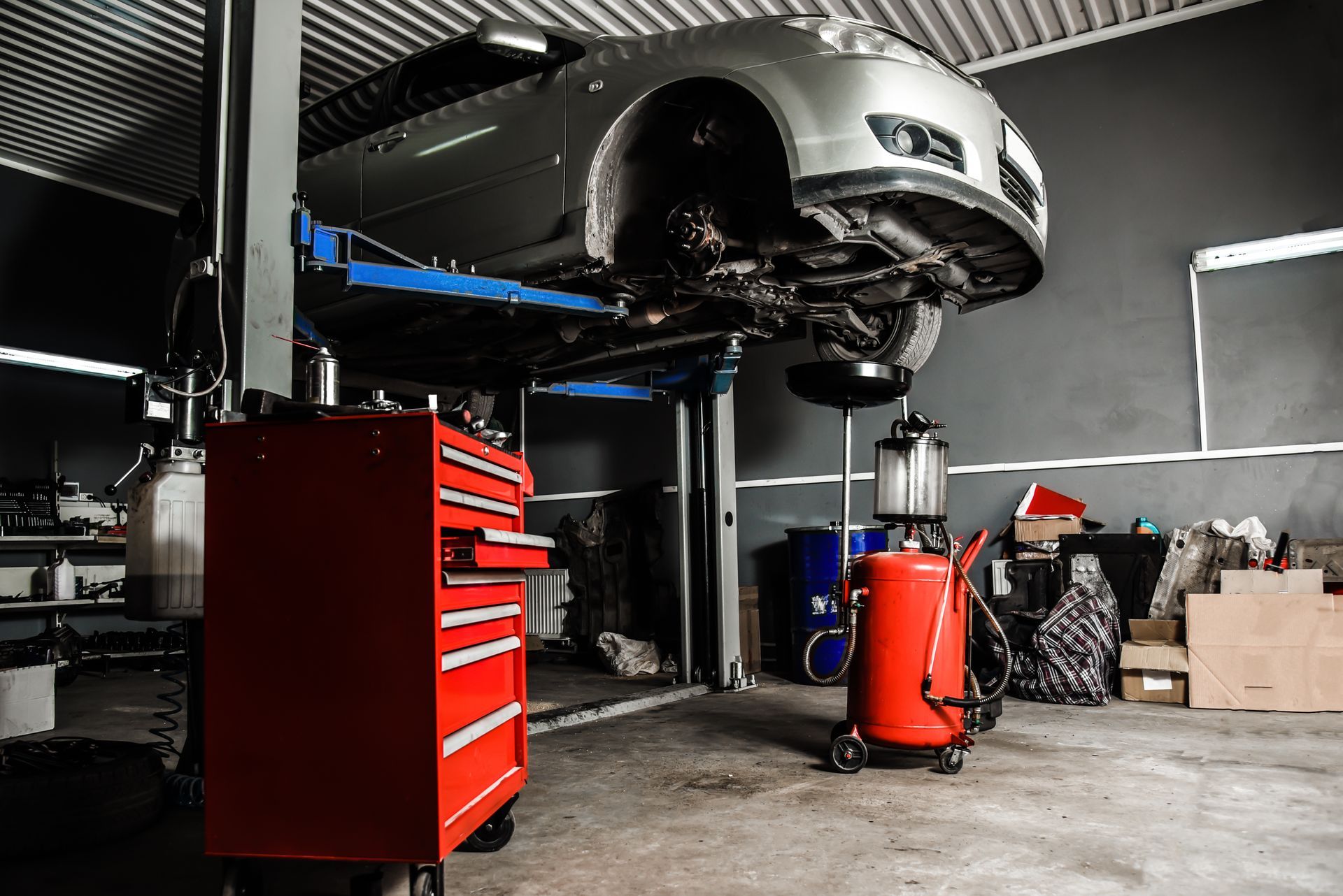 Car on a lift in a repair shop. Red toolbox and oil drain nearby.