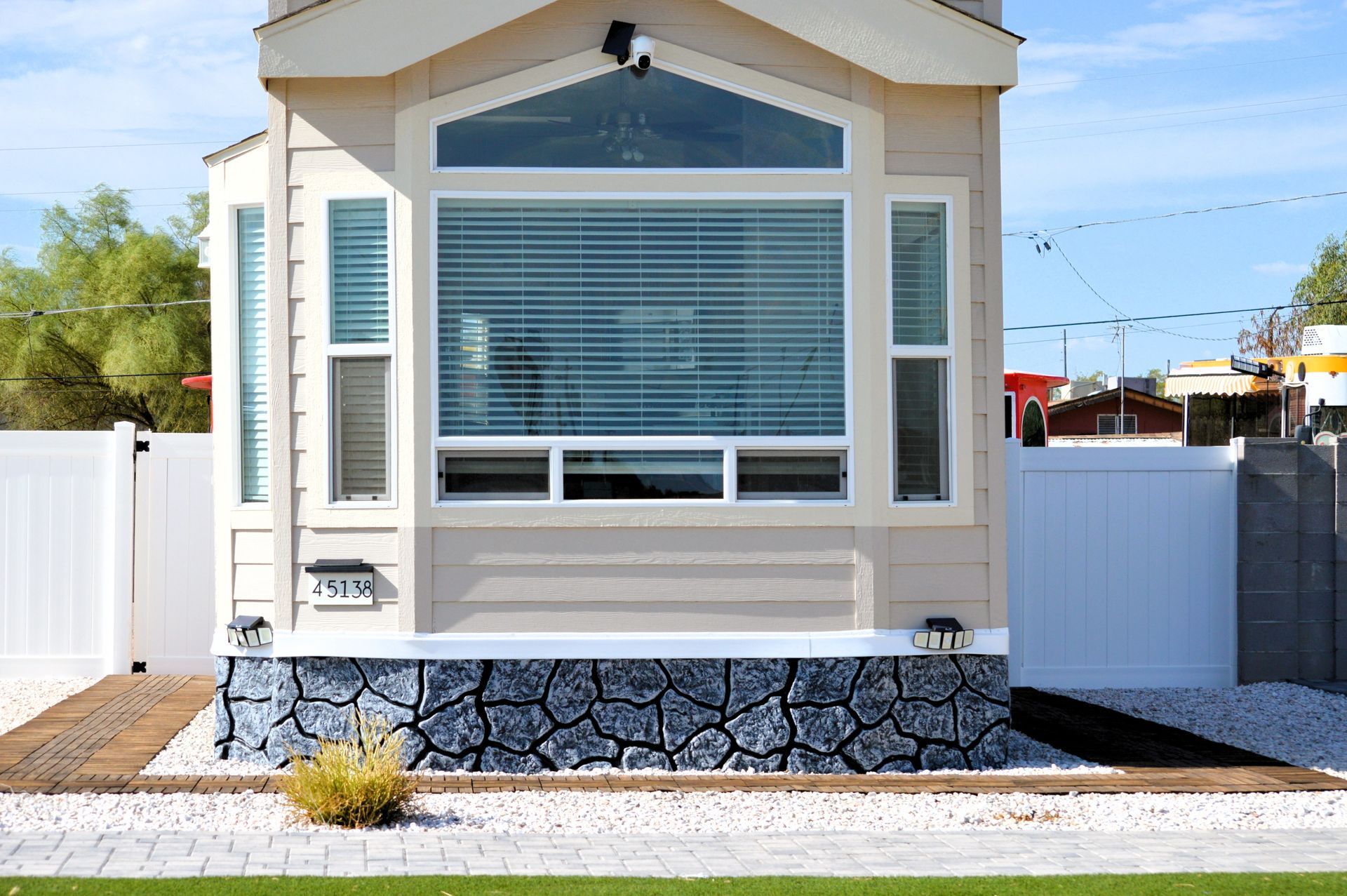 Small beige house exterior with large window, white fence, and stone skirting by the street