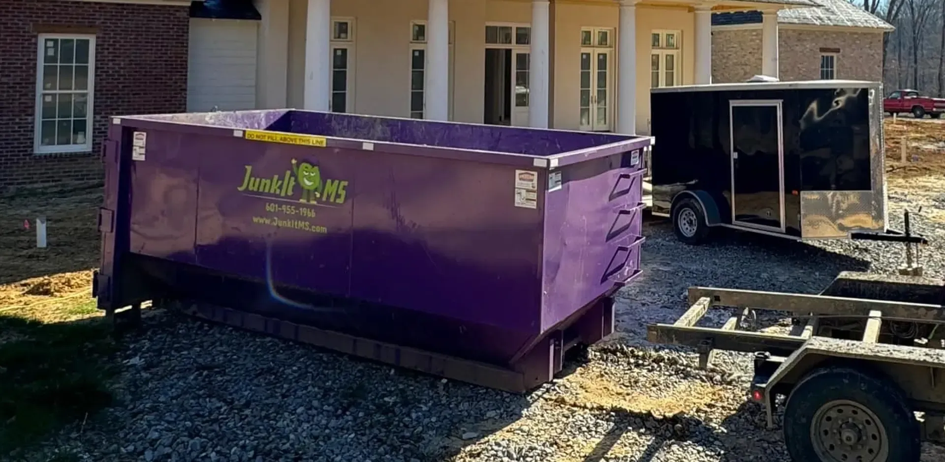 A purple dumpster in front of a house under construction, next to a black trailer.