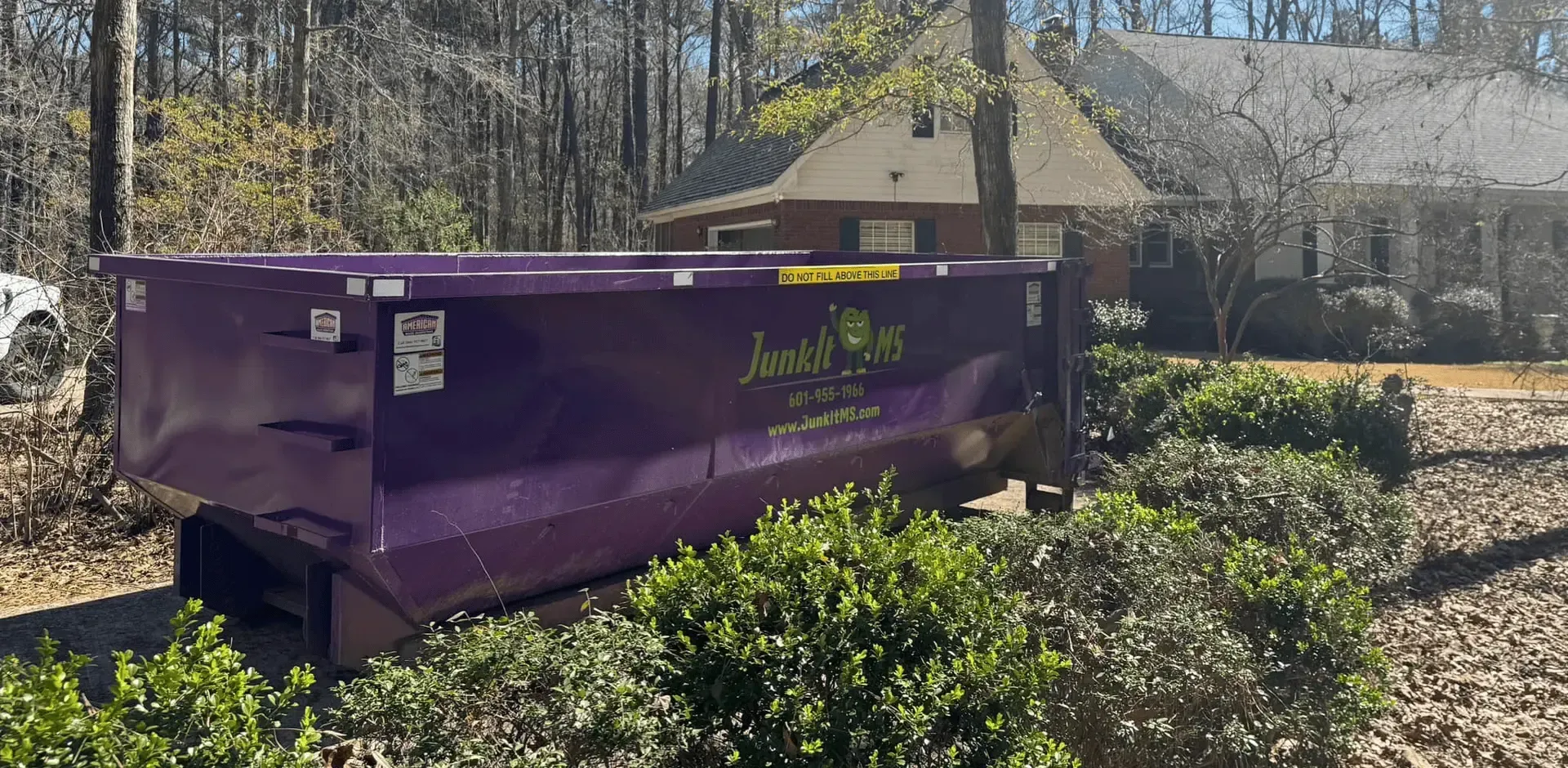 A purple dumpster in front of a house, surrounded by bushes, trees, and blue sky.