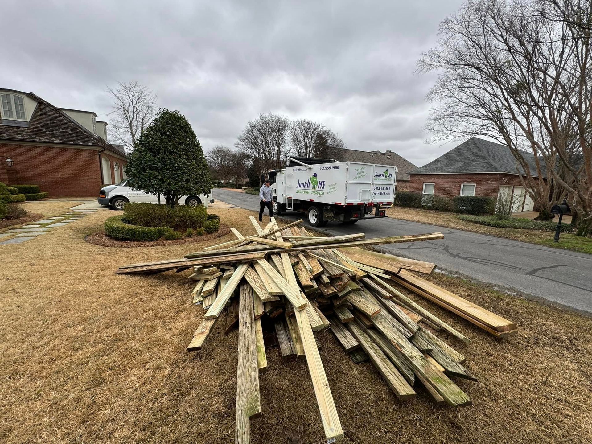 Pile of wooden boards in front of a yard, truck in the background, residential area.
