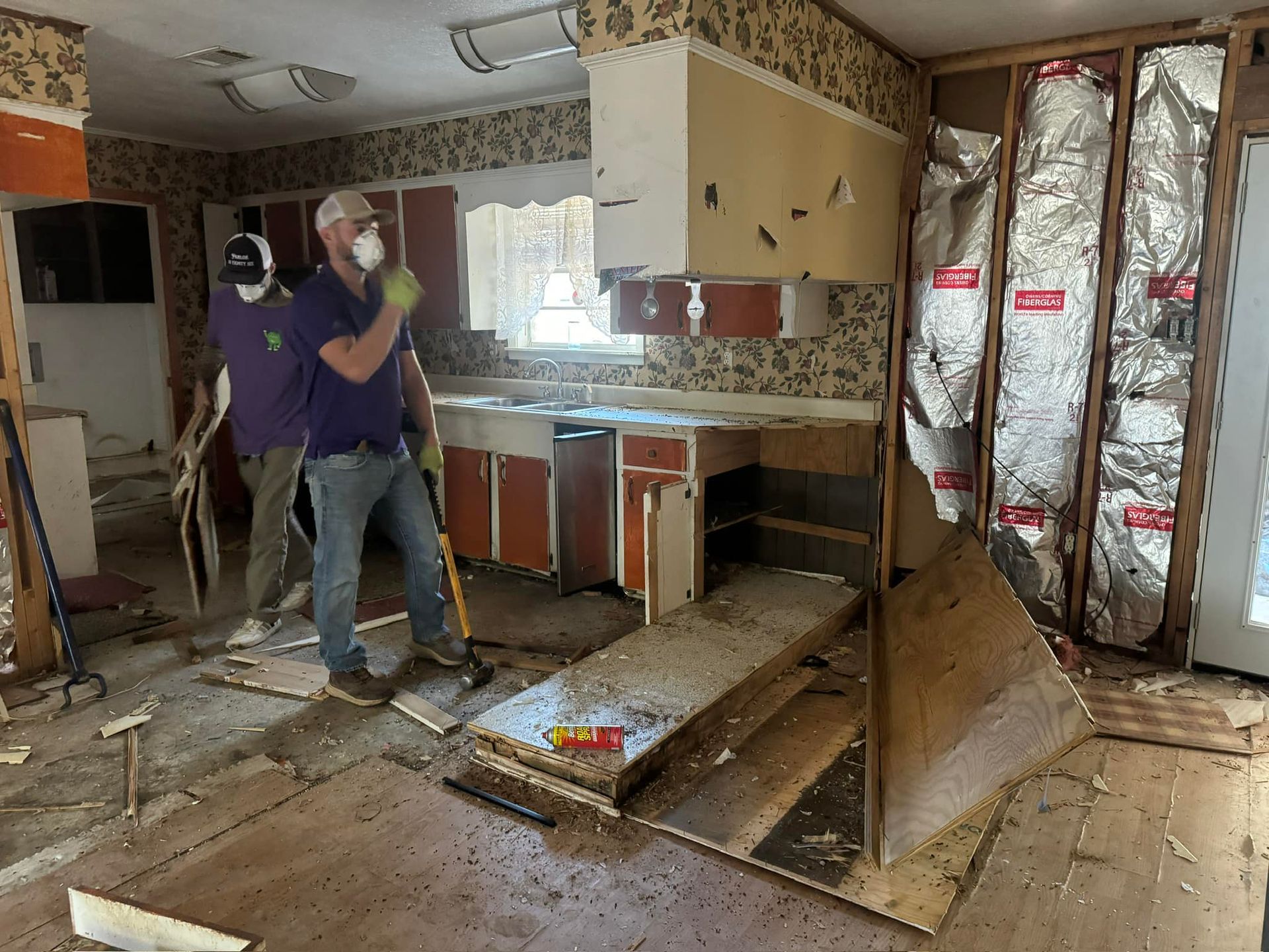 Two people demolishing a kitchen, walls torn open, cabinets visible, debris on the floor.