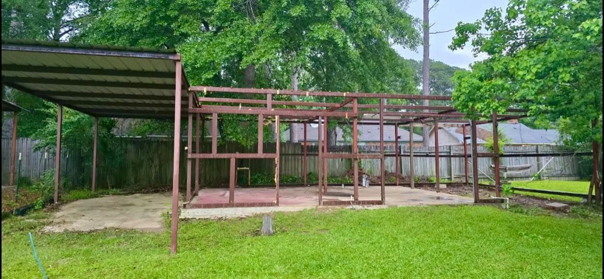 Metal structure frame in backyard, with a covered carport and wooden framework. Green lawn, trees, and fence.
