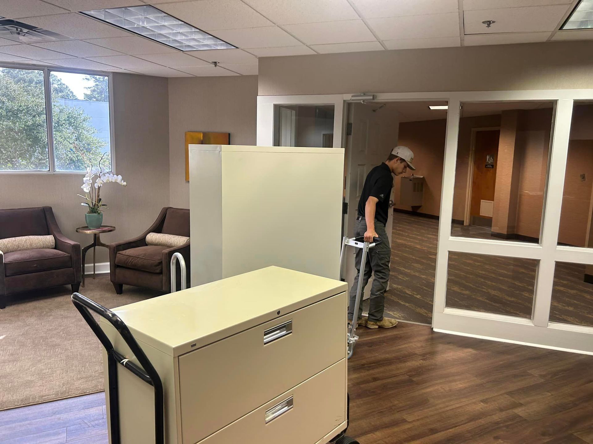 Person moving a beige file cabinet on a cart through an office doorway. There are other office furniture and another cabinet.