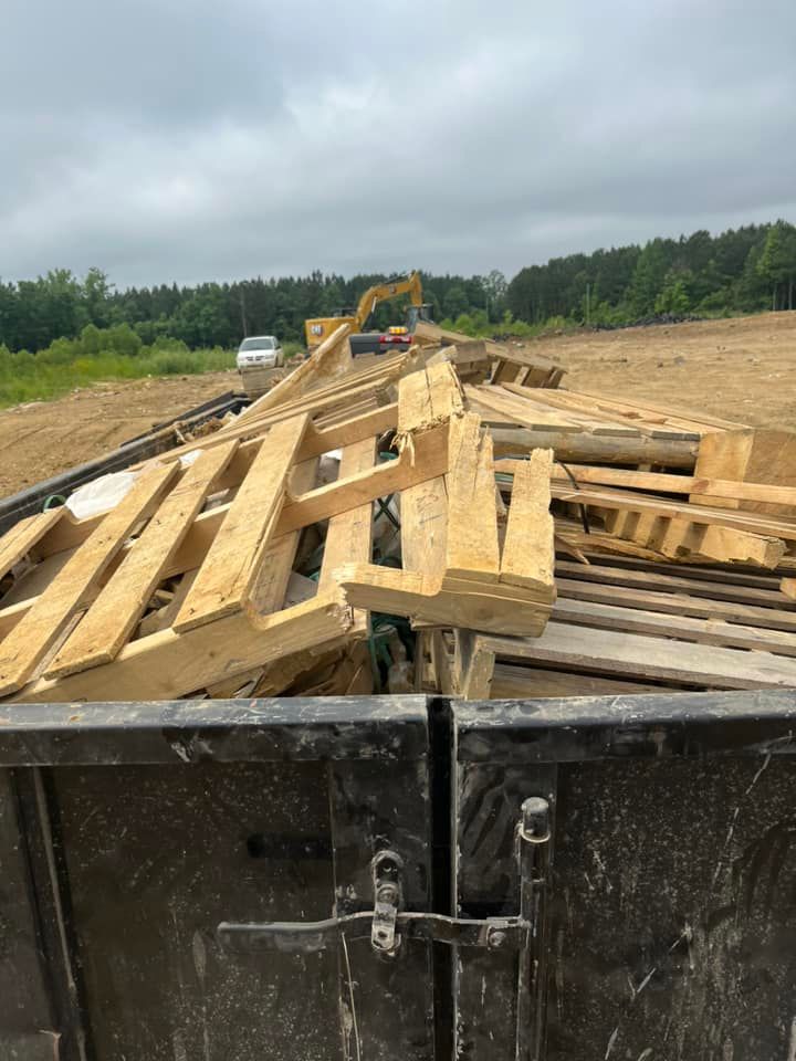 Wooden pallets piled high in a dumpster; construction site in the background with excavator.
