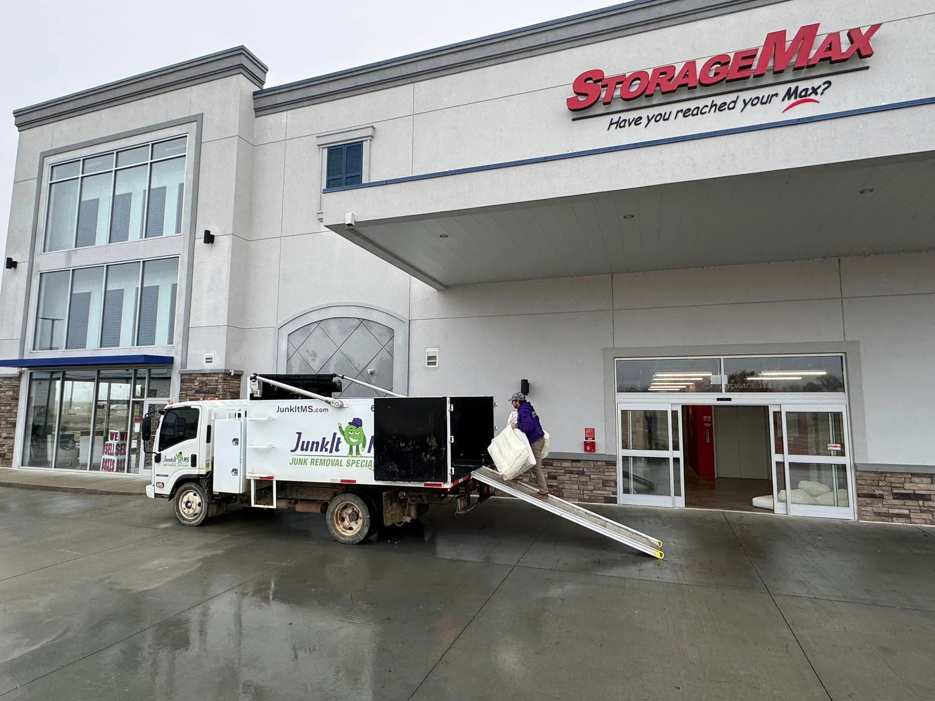 Truck loading into StorageMax building. Man in purple shirt. Cloudy day.